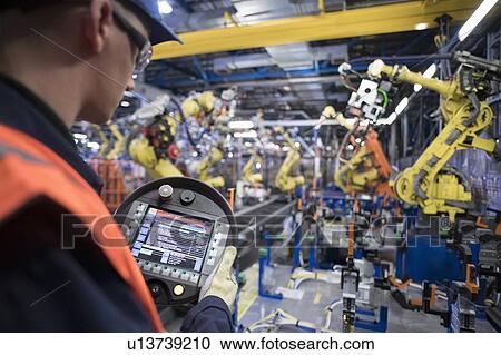 Stock Image - Apprentice engineer programming robots in car factory. Fotosearch