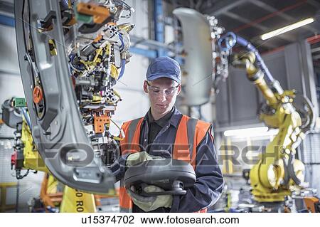 Stock Image - Apprentice engineer programming robots in car factory. Fotosearch