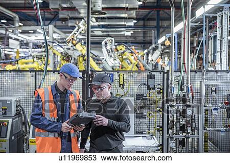 Stock Image - Apprentice engineer with mentor programming robots in car factory. Fotosearch