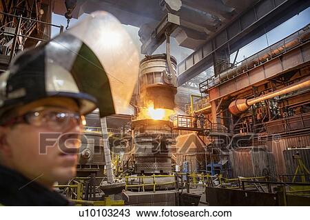 Stock Image - Apprentice steelworker in foreground during molten steel pour in steelworks. Fotosearch