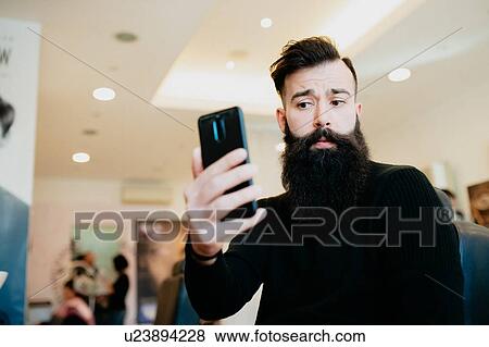 Stock Photo - Bearded man taking selfie in barber shop. Fotosearch