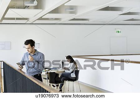 Stock Image - Businessman using smartphone in office corridor, colleague reading in background. Fotosearch