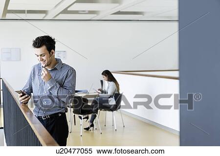Stock Photography - Businessman using smartphone in office corridor, colleague reading in background. Fotosearch