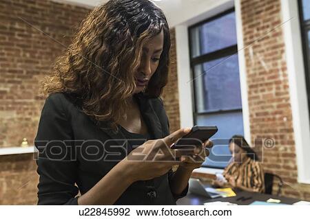 Stock Image - Businesswoman texting, colleague using laptop in background. Fotosearch