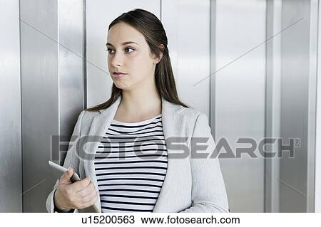 Stock Image - Businesswoman with digital tablet leaning against elevator door. Fotosearch