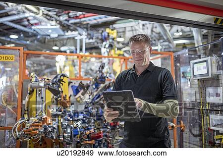 Stock Photo - Computer programmer using digital tablet with robots in car factory. Fotosearch