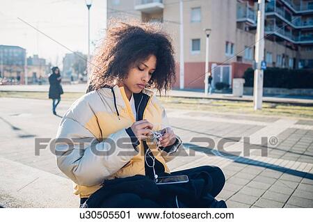 Cool young woman sitting on urban wall untangling earphones View Large Photo Image Stock Image - Cool young woman sitting on urban wall untangling earphones. Fotosearch