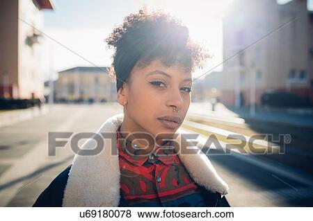 Stock Photo - Cool young woman with nose piercing on sunlit urban sidewalk, head and shoulder portrait. Fotosearch
