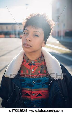 Stock Photography - Cool young woman with nose piercing on sunlit urban sidewalk, portrait. Fotosearch