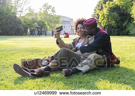 Stock Photo - Couple sitting on grass taking selfie, Milan, Italy. Fotosearch