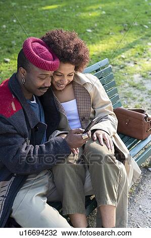Stock Image - Couple using cellphone on park bench, Milan, Italy. Fotosearch