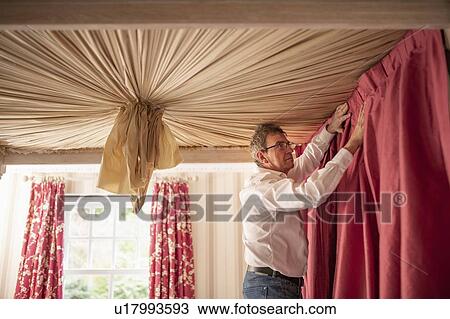 Stock Image - Curtain fitter fitting drapes to four poster bed. Fotosearch