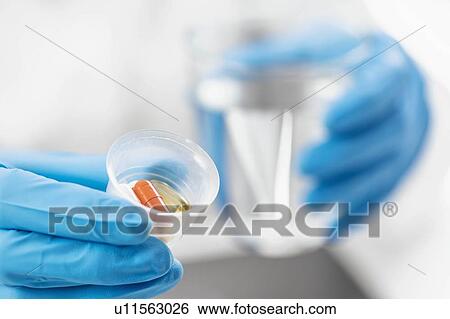 Stock Photograph - Doctor's hand in a glove holding graduated cup with pills. Fotosearch