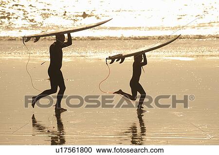 Stock Image - Father and son carrying surfboards on beach at sunset. Fotosearch