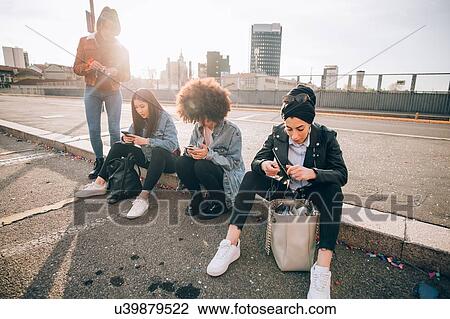 Stock Image - Friends sitting on kerb texting, Milan, Italy. Fotosearch