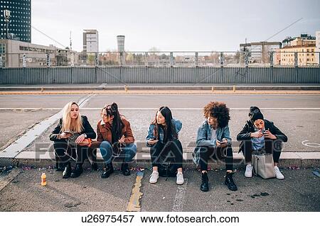 Friends sitting on kerb using smartphone, Milan, Italy View Large Photo Image Stock Photo - Friends sitting on kerb using smartphone, Milan, Italy. Fotosearch