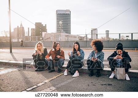 Stock Photo - Friends sitting on kerb using smartphone, Milan, Italy. Fotosearch