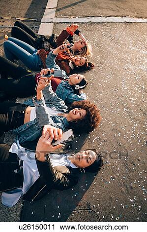 Stock Image - Friends together on kerb taking selfie and using smartphones, Milan, Italy. Fotosearch