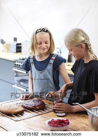 Stock Image - Girl and her sister baking a cake, smoothing jam onto cake at kitchen table. Fotosearch