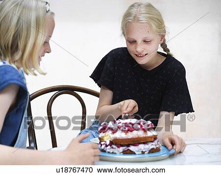 Stock Image - Girl and her sister slicing their homemade cake with fresh raspberries at kitchen table. Fotosearch
