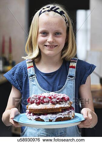 Stock Photograph - Girl baking a cake, holding homemade cake with raspberries on top in kitchen, portrait. Fotosearch