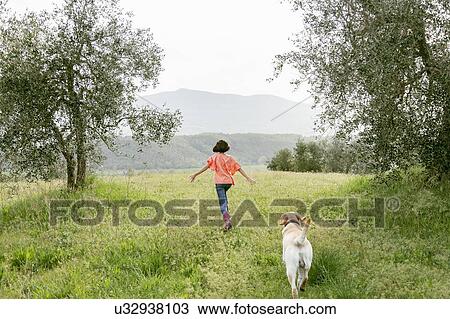 Stock Image - Girl running with labrador dog in scenic field landscape, rear view, Citta della Pieve, Umbria, Italy. Fotosearch