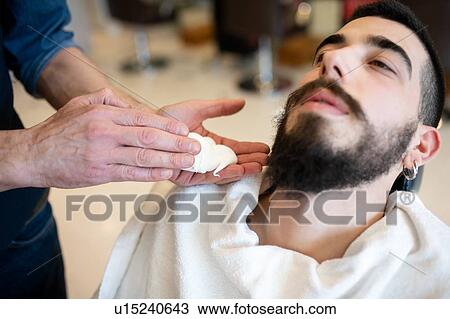 Stock Image - Hairdresser preparing to shave customer's beard in barber shop. Fotosearch