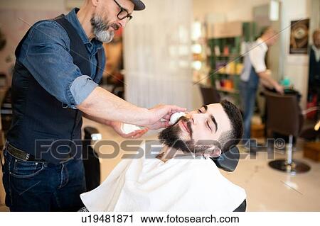 Stock Image - Hairdresser preparing to shave customer's beard in barber shop. Fotosearch
