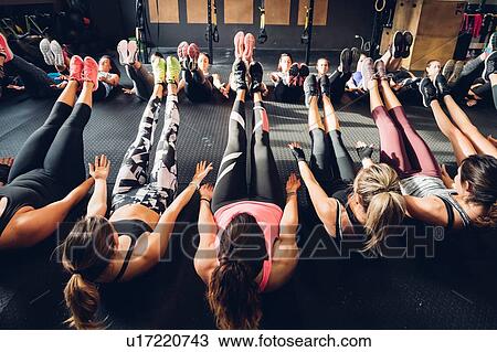 Stock Image - Large group of women training in gym, sitting on floor with legs raised. Fotosearch