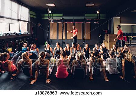 Stock Image - Large group of women training in gym with male trainers, sitting on floor. Fotosearch