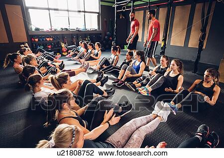 Stock Image - Large group of women training in gym with male trainers, sitting in rows on floor with legs raised. Fotosearch