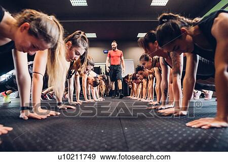 Stock Photo - Large group of women training in gym with male trainer, in rows doing push ups. Fotosearch