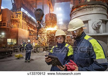 Stock Image - Male and female steelworkers using digital tablet during steel pour in steelworks. Fotosearch