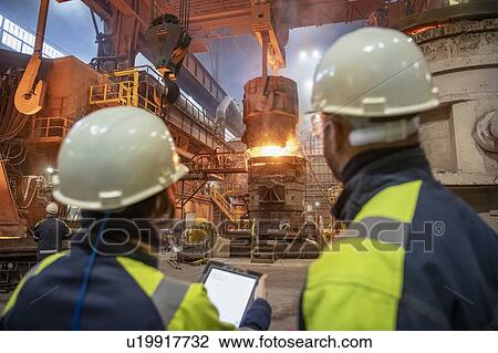 Stock Image - Male and female steelworkers using digital tablet during steel pour in steelworks. Fotosearch