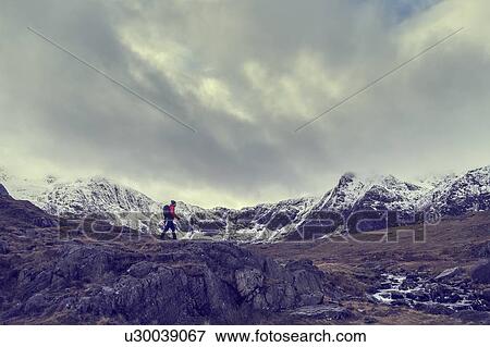 Stock Photo - Male hiker hiking across rugged landscape with snow capped mountains, Llanberis, Gwynedd, Wales. Fotosearch