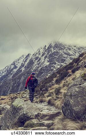 Stock Image - Male hiker hiking in rugged landscape with snow capped mountains, Llanberis, Gwynedd, Wales. Fotosearch