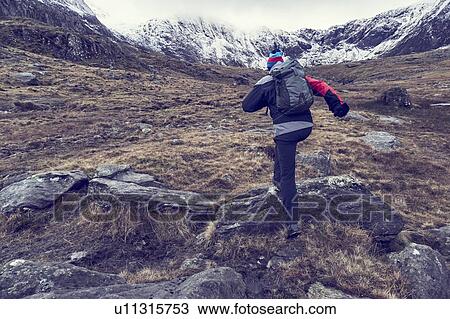 Stock Image - Male hiker hiking up rugged landscape with snow capped mountains, rear view, Llanberis, Gwynedd, Wales. Fotosearch