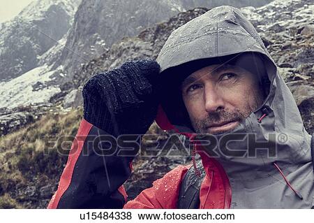 Stock Photo - Male hiker pulling hood up in sleeting snow capped mountain landscape, close up, Llanberis, Gwynedd, Wales. Fotosearch