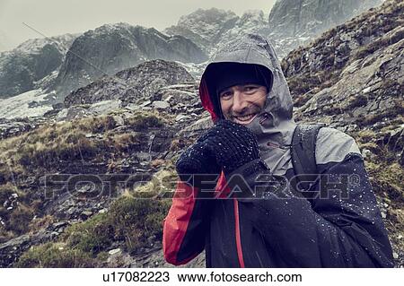 Stock Image - Male hiker with hood up in sleeting snow capped mountain landscape, portrait, Llanberis, Gwynedd, Wales. Fotosearch