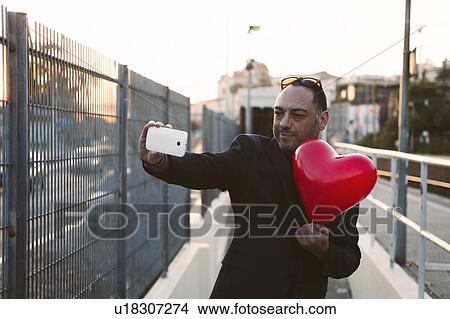 Picture - Man taking selfie with red heart shaped balloon. Fotosearch