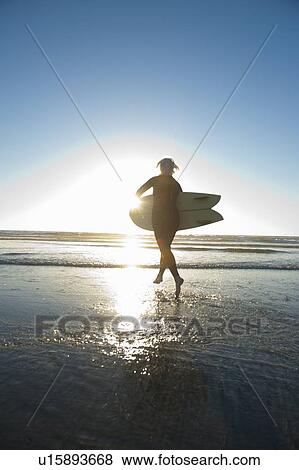 Stock Photo - Mature woman carrying surfboard into sea at sunset. Fotosearch
