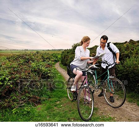 Stock Image - Mid adult cycling couple on rural dirt track looking at map, Southwold, Suffolk, UK. Fotosearch