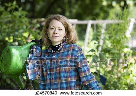 Stock Image - Mid adult woman carrying watering can in her garden. Fotosearch