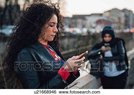 Stock Photograph - Mid adult woman with long curly hair looking at smartphone by city canal. Fotosearch