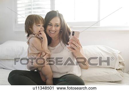 Stock Image - Mother and daughter taking selfie on bed. Fotosearch