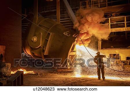 Stock Image - Steelworker cleaning pouring end of flask during steel pour in steelworks. Fotosearch