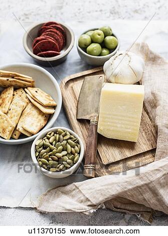 Stock Photography - Still life of cutting board and cheese with bowls of crackers, olives, salami and pumpkin seeds, overhead view. Fotosearch