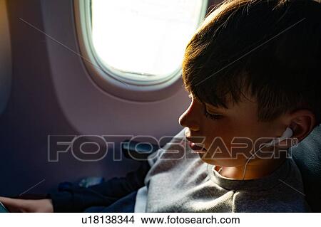 Picture - Teenage boy listening to music on earphones during airplane journey, close up. Fotosearch