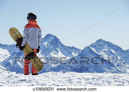 Stock Image - Teenage boy snowboarder looking out over landscape from snow covered mountain top, rear view, Alpe-d'Huez, Rhone-Alpes, France. Fotosearch