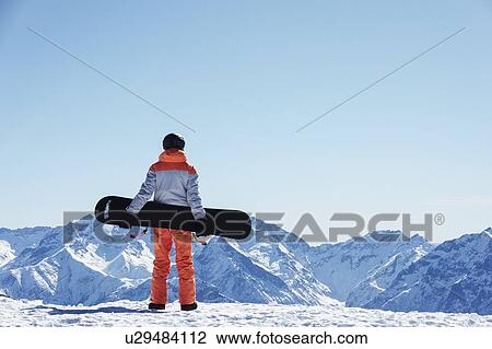 Stock Image - Teenage boy snowboarder looking out over landscape from snow covered mountain top, rear view, Alpe-d'Huez, Rhone-Alpes, France. Fotosearch
